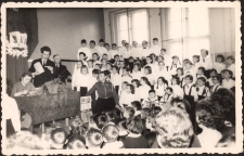 Chór Szkoły Podstawowej nr 1 w sali gimnastycznej, ok. 1965 r. - przy stole prezydialnym pierwsza z lewej Jadwiga Kielar, trzeci dyrektor szkoły Zieliński [Dokument ikonograficzny]
